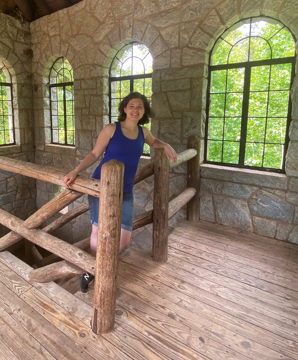 Rebecca stands at the top of wooden stairs with windows behind her