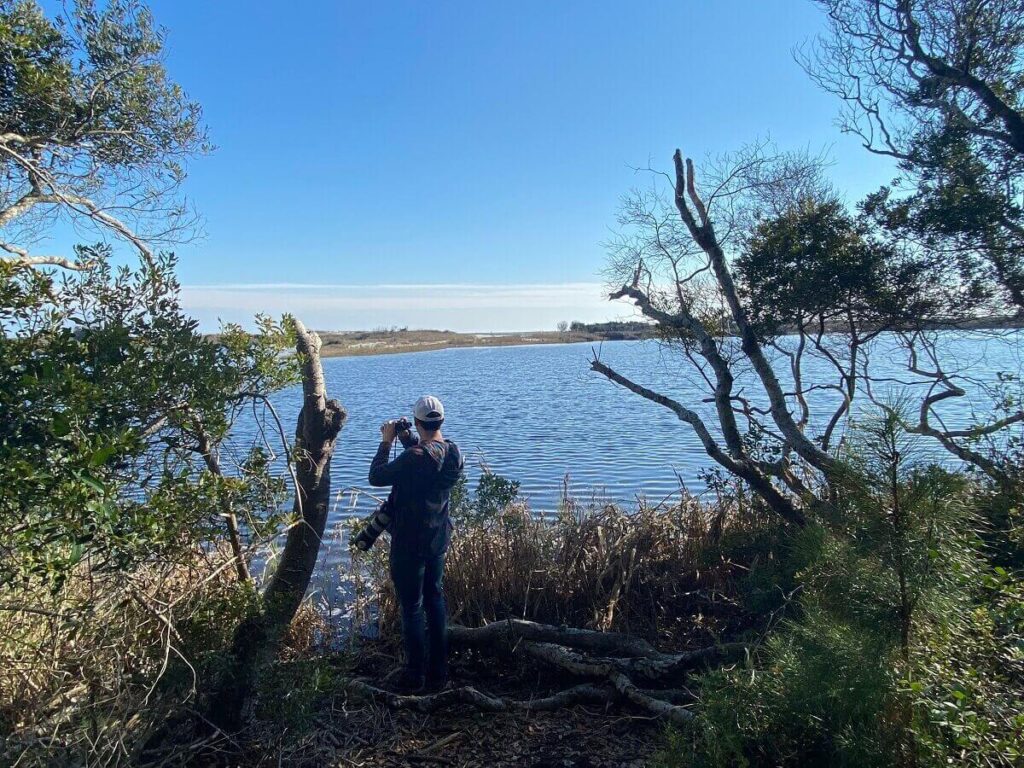 A man with binoculars and a camera looks out at a coastal pond