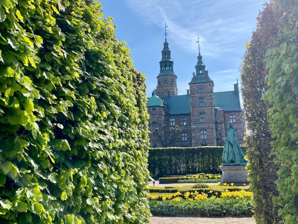 Partial view of Rosenborg Castle through two tall hedges with a statue of a Danish queen